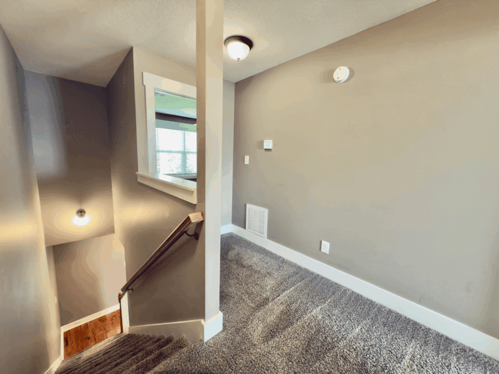 A carpeted stairway landing with beige walls, a wooden handrail, a wall vent, thermostat, smoke detector, and a window with blinds.