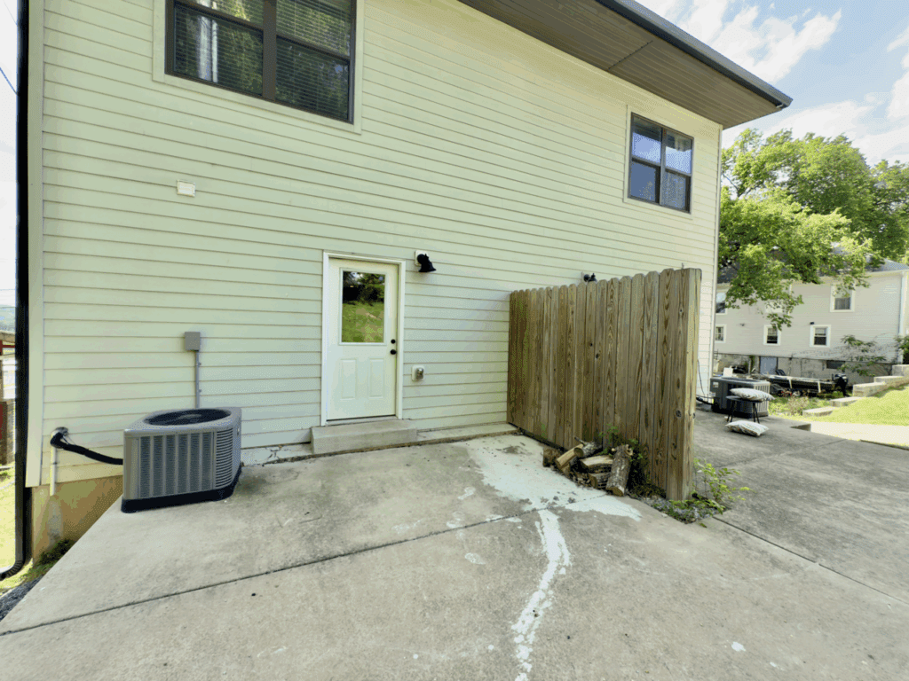 Back view of a house with a white door, central AC unit, wooden privacy fence, and concrete patio. Neighboring houses and some outdoor items are visible in the background.