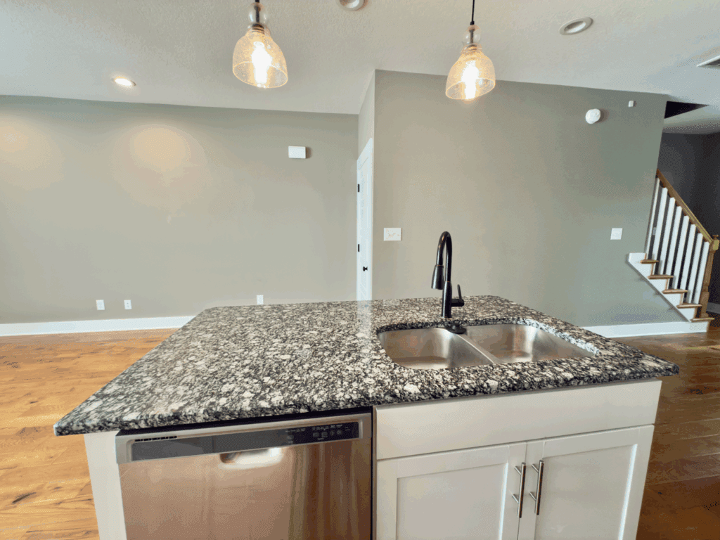 A modern kitchen island with a granite countertop, stainless steel dishwasher, double sink, and black faucet; hardwood floors and stairs are visible in the background.