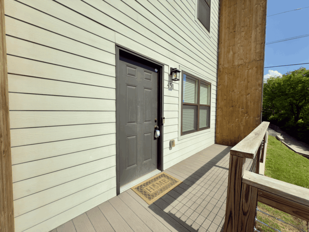 A gray front door with a lockbox, house number 451, welcome mat, and two windows, set on a wooden porch with a railing.