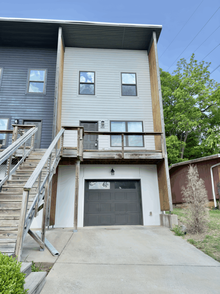Modern two-story townhouse with gray siding, a wooden balcony, exterior stairs, and a closed garage door, next to a red shed and greenery.