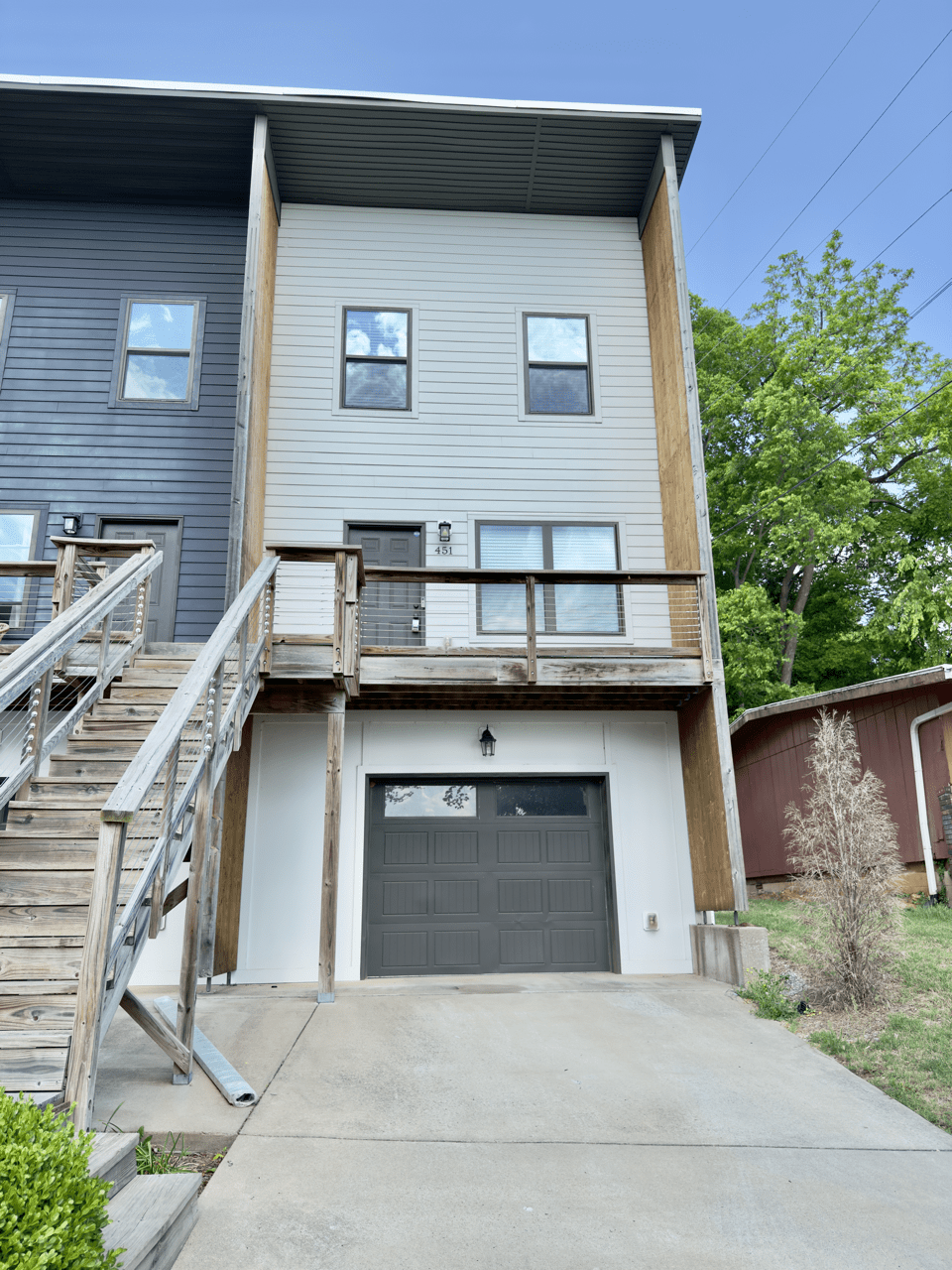 Modern two-story townhouse with gray siding, a wooden balcony, exterior stairs, and a closed garage door, next to a red shed and greenery.